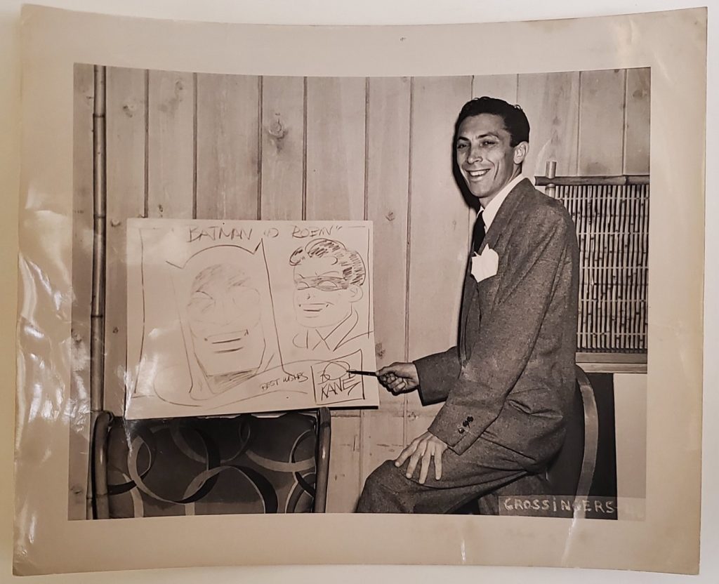 Bob Kane pointing to a whiteboard illustration of Batman